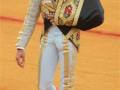 Un torero en traje tradicional de color blanco y negro, con detalles dorados y flores rojas. Lleva una falda larga blanca, guantes negros y calcetines rosados. El fondo es un campo de lucha de arena roja.