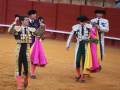 Tres toreros caminan en una plaza de toros, cada uno con un capote colorido y un sombrero tradicional. El suelo es de tierra, y las paredes rojas del coso están al fondo.