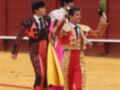 Un torero en traje de luces y otro en traje de cuero caminan con orgullo por la plaza de toros, sosteniendo un ramo de flores.