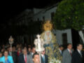 La procesión nocturna muestra una imagen de la Virgen con un manto dorado y una corona, rodeada por fieles en trajes formales. La escena se desarrolla frente a un edificio blanco con balcones, iluminada por faroles.