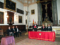 Un grupo de personas sentadas en una sala histórica, rodeadas por cuadros y un crucifijo. La mesa central está cubierta con una mantilla roja, y hay un crucifijo en el fondo.