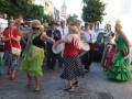 Fotografía de una celebración en un pueblo, con personas bailando y vestidas con trajes tradicionales. En el fondo, se puede ver una iglesia con un campanario y coches estacionados.