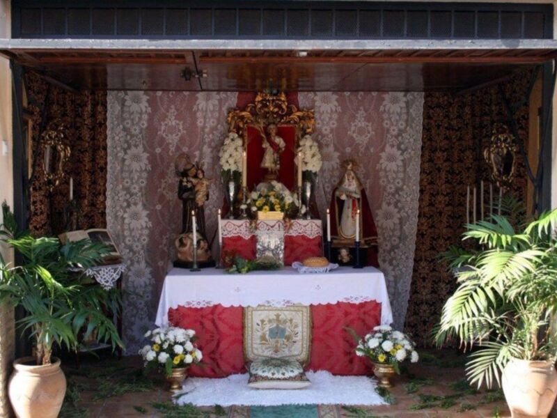 Un altar religioso con estatuas de San Antonio de Padua y San José, adornado con flores y velas, en un ambiente con cortinas de encaje y plantas verdes.