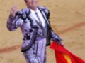 Un torero en traje tradicional de color blanco y negro con detalles dorados, sosteniendo una capa roja en la mano izquierda. El fondo es un terreno de toros de color arena.