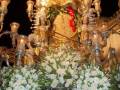 Imagen de una Virgen María en procesión, rodeada de flores blancas y luces doradas.