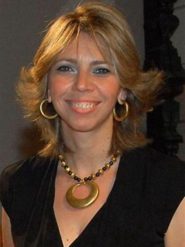 A una mujer con cabello rubio largo y ondulado, llevando un vestido negro y un collar de cuentas doradas con una gran medalla circular.