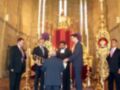Una ceremonia religiosa en una iglesia con un altar central, velas encendidas y una figura dorada. Un hombre en traje negro se inclina para besar el pie de otro hombre en traje azul. Otros hombres observan la ceremonia, con un altar dorado y una cruz en el fondo.