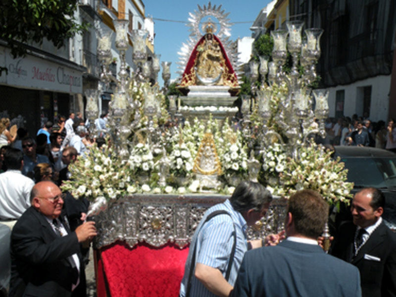 Imagen de una procesión religiosa con una imagen venerada en un carro decorado con flores y luces, rodeada por fieles observando la celebración.