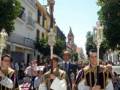 Una procesión religiosa en un entorno urbano. Personas vestidas con trajes tradicionales y capas bordadas participan en la procesión, seguidas por una figura crucificada. El cielo es claro y el ambiente parece ser de un día soleado, con edificios y árboles al fondo.