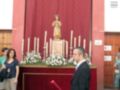 Una persona en traje negro sostiene un bastón rojo, frente a una estatua religiosa con velas y flores en un altar rojo. Detrás, una gran cortina roja cubre la pared de un edificio con puertas y ventanas.