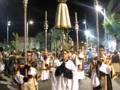 Procesión nocturna con sacerdotes en trajes tradicionales portando cruces y estandartes, en una calle iluminada.