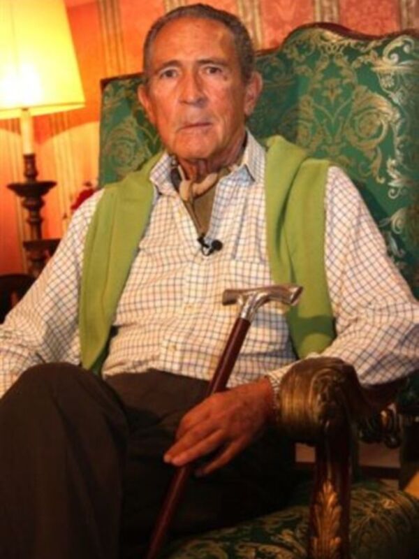 Un hombre mayor con una camisa a cuadros, un chaleco verde y un bastón. Está sentado en una silla con un fondo de cortinas coloridas.