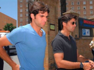 Dos hombres jóvenes caminan por una calle con edificios a ambos lados. El hombre de la izquierda lleva una camiseta azul y tiene el brazo en la cadera. El hombre de la derecha lleva una camiseta negra y un reloj en el brazo. En el fondo, se puede ver una agencia de seguros y un coche estacionado.
