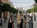 Fotografía de una procesión en un pueblo, con personas vestidas formalmente y portando banderas. La imagen muestra una calle con casas blancas y verdes, adornadas con guirnaldas. La gente camina en fila, algunas personas llevan banderas y otros objetos ceremoniales.