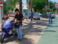 Un hombre en una camiseta negra y pantalones azules está preparando su casco de motocicleta en un aparcamiento. Hay varios coches y una moto en el fondo, con palmeras y edificios a lo lejos.