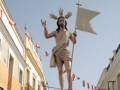 Imagen de una procesión religiosa con un Cristo resucitado en alto, portando una bandera blanca y rodeado de flores. Fondo de edificios con banderas ondeando en el aire.