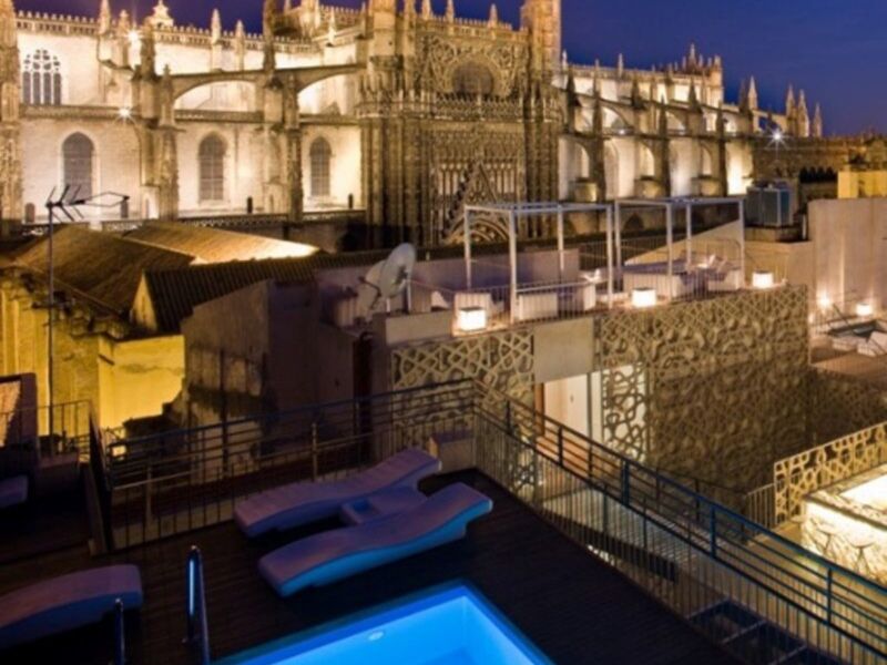 Una piscina con dos sillones azules en un terraplén, frente a la catedral de Sevilla iluminada por la noche. La piscina está rodeada por una estructura metálica con luces que reflejan en el agua. La catedral, con su arquitectura gótica, se destaca en el fondo.