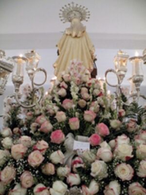 Una estatua de la Virgen María rodeada por una gran composición floral con rosas y otros tipos de flores, en un entorno decorado con candelabros.