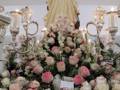 Una estatua de la Virgen María rodeada por una gran composición floral con rosas y otros tipos de flores, en un entorno decorado con candelabros.