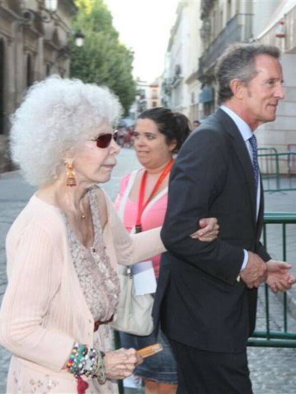 Una mujer con cabello blanco y gafas de sol, posiblemente en una celebración o evento, camina junto a un hombre en traje formal. La escena parece ser en una calle de ciudad, con edificios y árboles al fondo.