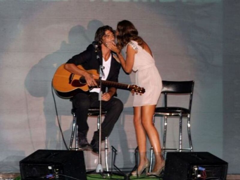 Un hombre y una mujer están en un escenario, el hombre toca la guitarra mientras la mujer le canta. Ambos están sentados en sillas, con un altavoz y una pantalla de proyección detrás de ellos.