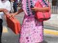 Una mujer en un vestido rosa con flores, caminando por una calle con un bolso rosa y un bolso con rayas rojas y blancas.