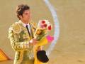 Un torero en traje tradicional sostiene un ramo de flores mientras camina por una arena amarilla con una línea blanca.