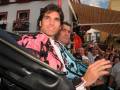 Dos hombres sonrientes en un evento, uno con chaqueta rosa y otro con camisa azul. El ambiente parece ser un evento público con espectadores en el fondo.