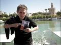 Un hombre sostiene un libro con la imagen de un hombre en la portada, frente a una vista panorámica de Sevilla, con el Torre del Oro y el río en el fondo.