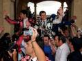 Un grupo de personas celebran en una entrada histórica, con un hombre en el centro levantado por los hombros. La gente está tomando fotos con cámaras de mano y otros dispositivos, mientras el ambiente es festivo.