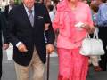 Elegante pareja caminando juntos por una calle, el hombre usa traje y corbata, la mujer lleva un vestido rosa y pantalones con estampado, ambos sonríen y parecen estar en una celebración.