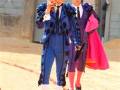 Dos toreros en trajes tradicionales, uno con sombrero y el otro con capa roja, caminan en un campo de toros.