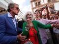 Una persona con una chaqueta verde y un vestido rojo está tomando una selfie en la calle, rodeada de personas que también están fotografiándose. La imagen está tomada en un entorno urbano con edificios y cielo azul.