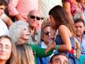 Una anciana con cabello blanco y una joven con vestido azul a cuadros están abrazándose en medio de un grupo de personas. La anciana lleva una pulsera y la joven tiene una corbata roja. El ambiente parece ser un evento público con gente de diferentes edades y estilos vestimentarios.