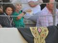 Una mujer en verde está sujetando un objeto blanco, mientras otras personas observan desde atrás una estructura con un tapiz bordado en negro y dorado.