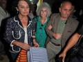 Una mujer con una chaqueta azul y un vestido de rayas camina mientras sostiene una bolsa. Detrás de ella, otra mujer con un abrigo verde y gafas de sol camina. Un hombre con una chaqueta marrón y una corbata negra también camina detrás. La escena parece ser nocturna en un lugar con iluminación artificial.