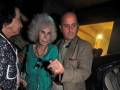 Una anciana con cabello blanco y una peluca azul está siendo ayudada a salir de un vehículo, seguida por un hombre en traje.