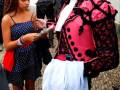 Un hombre en traje de carnaval rojo y negro con un chaleco blanco está firmemente firmando una tarjeta de visita para una mujer en un vestido azul con lunares. Ambos están en una calle de piedra, con otros individuos y edificios al fondo.