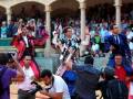 Un grupo de personas en trajes tradicionales se encuentra en un escenario rodeado por espectadores. La imagen captura una celebración o evento cultural, con el público sentado en gradas y algunos fotógrafos tomando fotos. La escena está ambientada en un entorno histórico o de tradición, con columnas y arquitectura que sugieren una plaza pública.