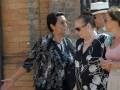 Las tres personas están caminando juntas, dos de ellas son mujeres mayores con vestidos oscuros y joyas, mientras que la tercera es un hombre con una gorra blanca. La escena parece ser en un entorno urbano, posiblemente una calle o pasillo de edificio.