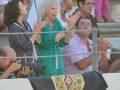 Un grupo de personas en una tribuna, algunas con cámaras y bebidas. La imagen muestra un detalle de la parte superior de una silla con un diseño floral.