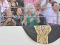 Una mujer con una camisa verde y un sombrero negro con detalles dorados está entre otros espectadores en un evento.