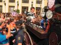 Un hombre vestido con traje de matador posa en el techo de un coche antiguo en medio de una multitud en una plaza.