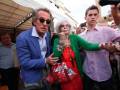 Una mujer con cabello blanco y gafas, vestida con rojo y verde, es acompañada por un hombre en azul y otro joven en rosa, en un evento público con otros espectadores alrededor.
