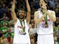 Dos jugadores de baloncesto celebran con trofeos y medallas, uno levantando un trofeo grande. El fondo muestra a una multitud de espectadores en un estadio lleno, con una camiseta visible que dice "España" y el año 2013.