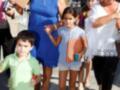 Una familia camina por un camino de tierra, con una mujer en el centro vestida con un vestido azul y dos niñas a su lado, una de ellas sosteniendo un instrumento musical.