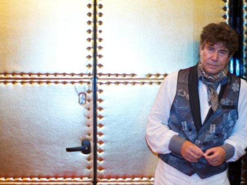 Un hombre con una camisa blanca, chaleco azul y bufanda de seda posa frente a una puerta con detalles dorados.