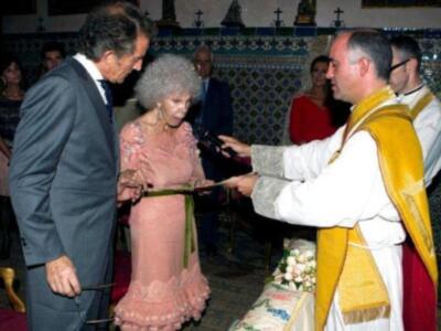 Una ceremonia religiosa en un ambiente tradicional, con una mujer vestida de rosa y un hombre en traje negro. Un sacerdote viste amarillo con una saya blanca, bendice a la mujer. La pared tiene azulejos de diseño geométrico y una imagen en el techo.