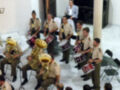 Una banda militar toca en un salón blanco, con instrumentos como tubas y tambores. Los músicos están vestidos con uniformes militares, y el ambiente es formal.