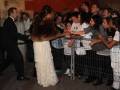 Una mujer en un vestido largo y con capa de piel se acerca a una fila de personas detrás de un cercado metálico. La escena parece ser en una alfombra roja o evento público nocturno, con la luz de la noche y la presencia de una multitud.